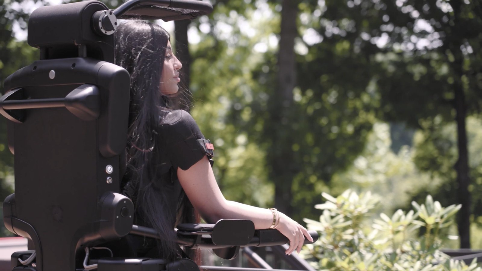 Une utilisatrice aux longs cheveux noirs, debout dans l’exosquelette Eve, regarde au loin sur une terrasse entourée d’arbres