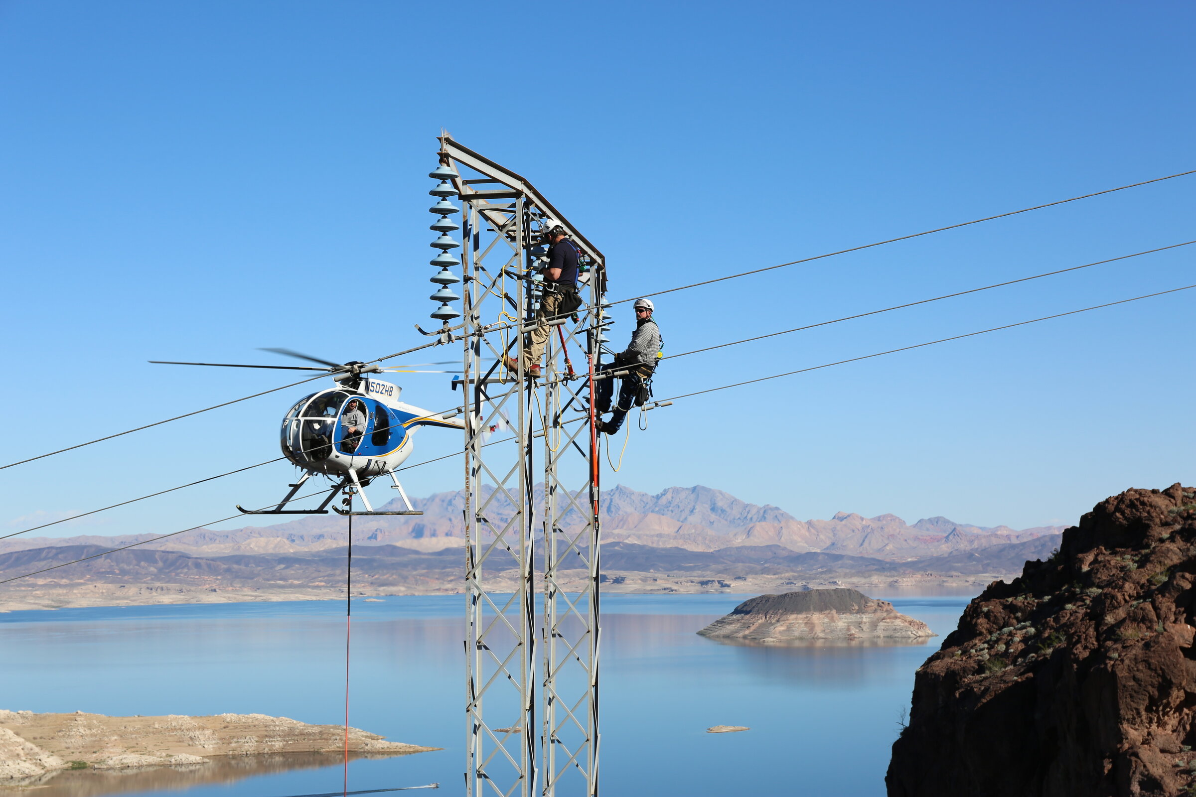 Deux monteurs en haut d’un pylône électrique au-dessus d’un lac, un hélicoptère en vol stationnaire à côté (photo d’illustration)