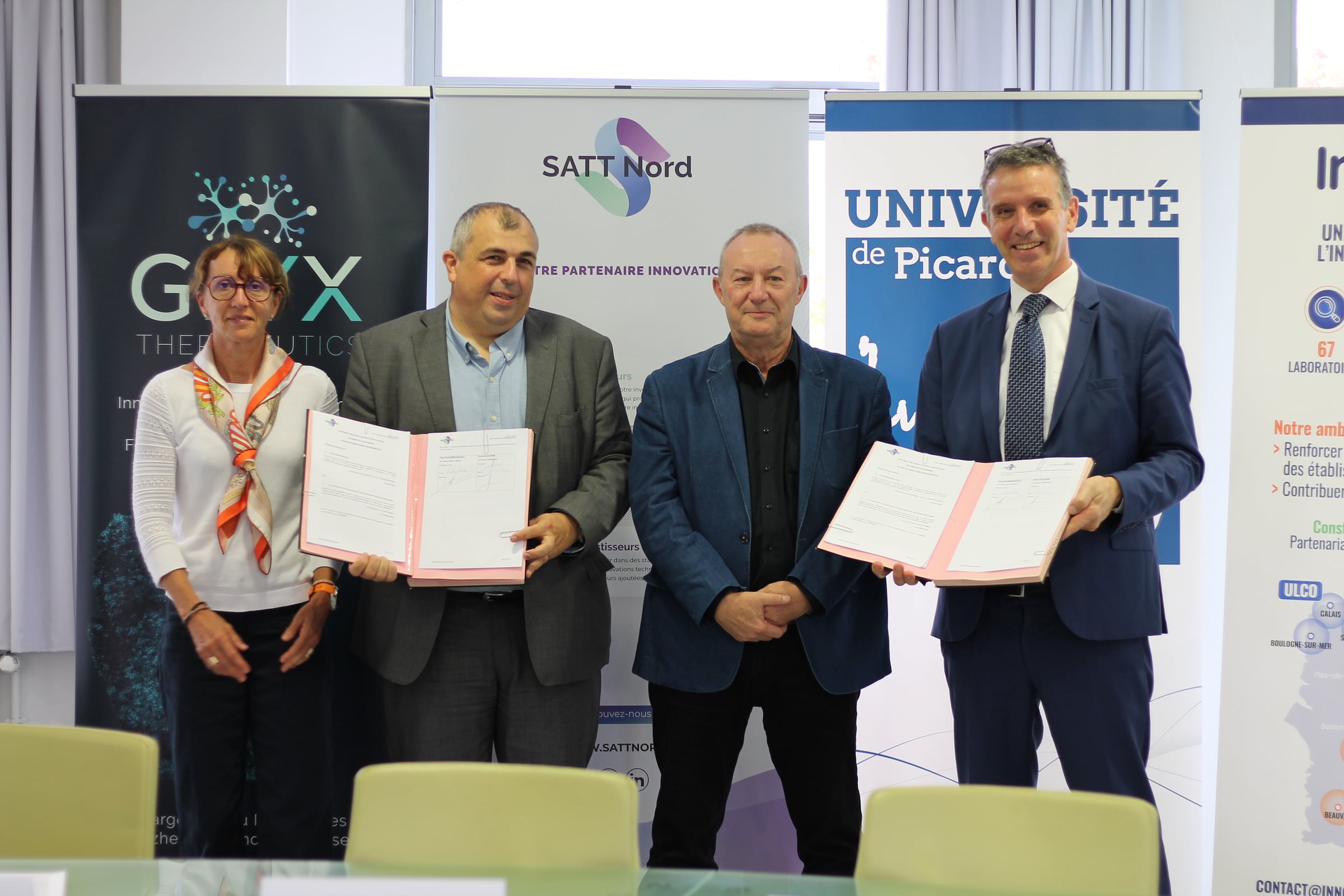 Quatre personnes souriantes derrière une table, deux d’entre elles brandissant des dossiers roses de la licence signée entre Glyx Therapeutics, la SATT Nord et l’UPJV