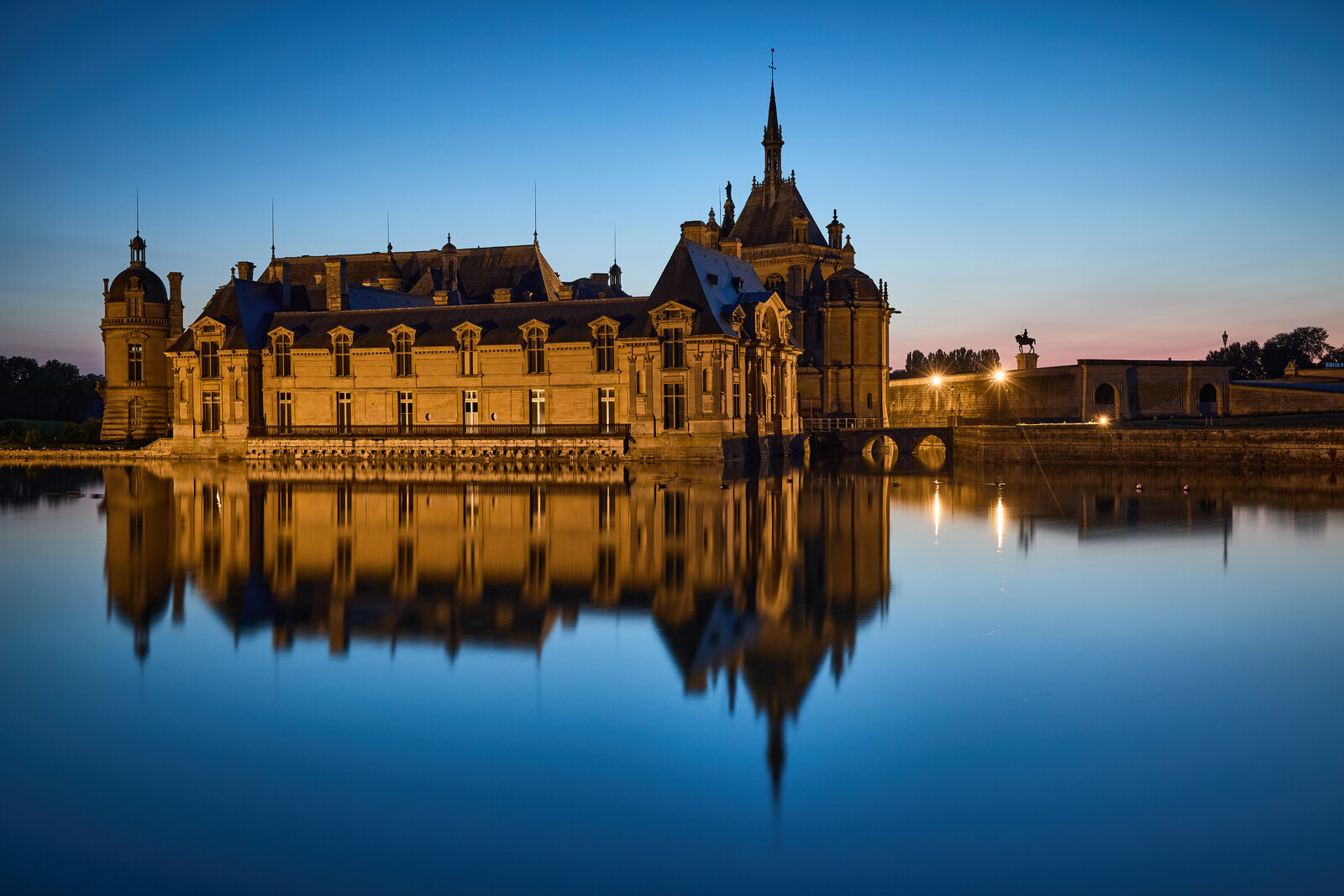Le château de Chantilly au lever du soleil, façades rose et pierre se reflétant dans les douves