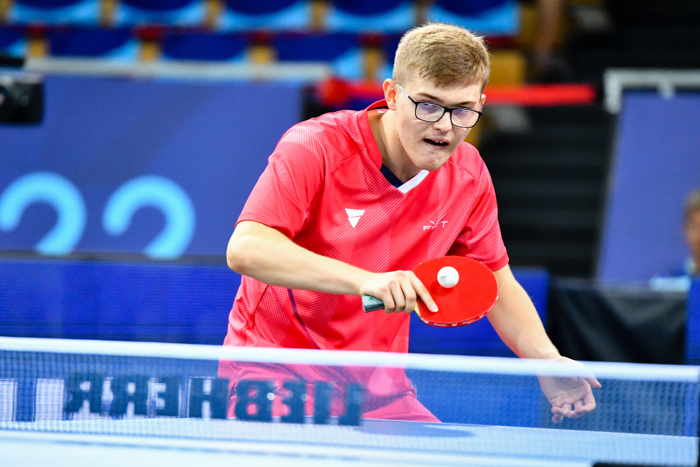 Alexis Lebrun en maillot rouge de l’équipe de France, concentré au-dessus de la table, raquette prête
