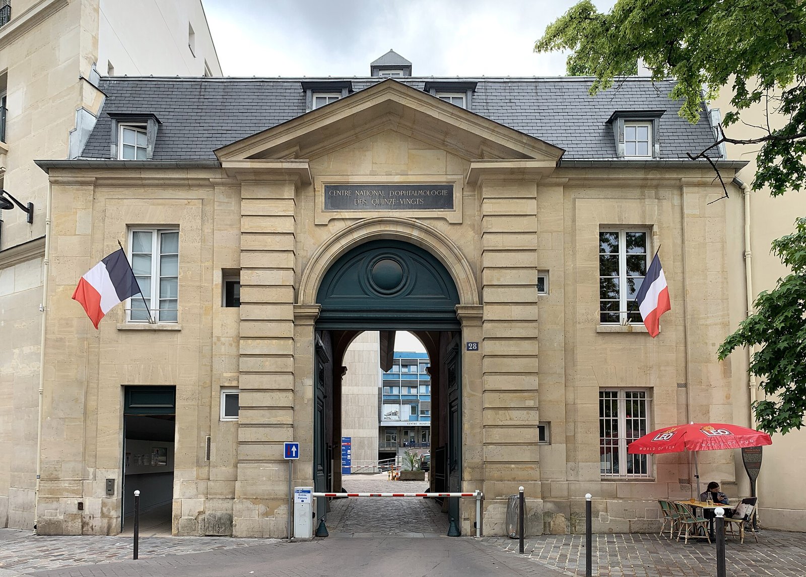 Le portail d’entrée de l’Hôpital national des Quinze-Vingts à Paris, encadré de drapeaux français, sous un ciel nuageux