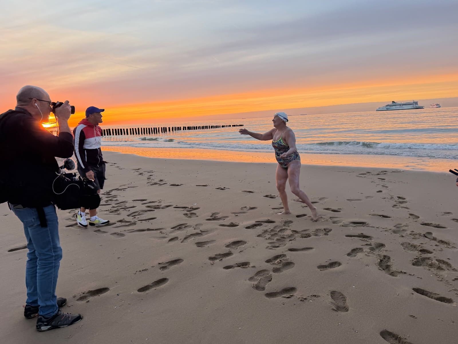 Salima Bouayad Agha, bonnet blanc et maillot de bain, tend les bras en sortant de l’eau sur la plage de Sangatte au coucher du soleil, face à un photographe