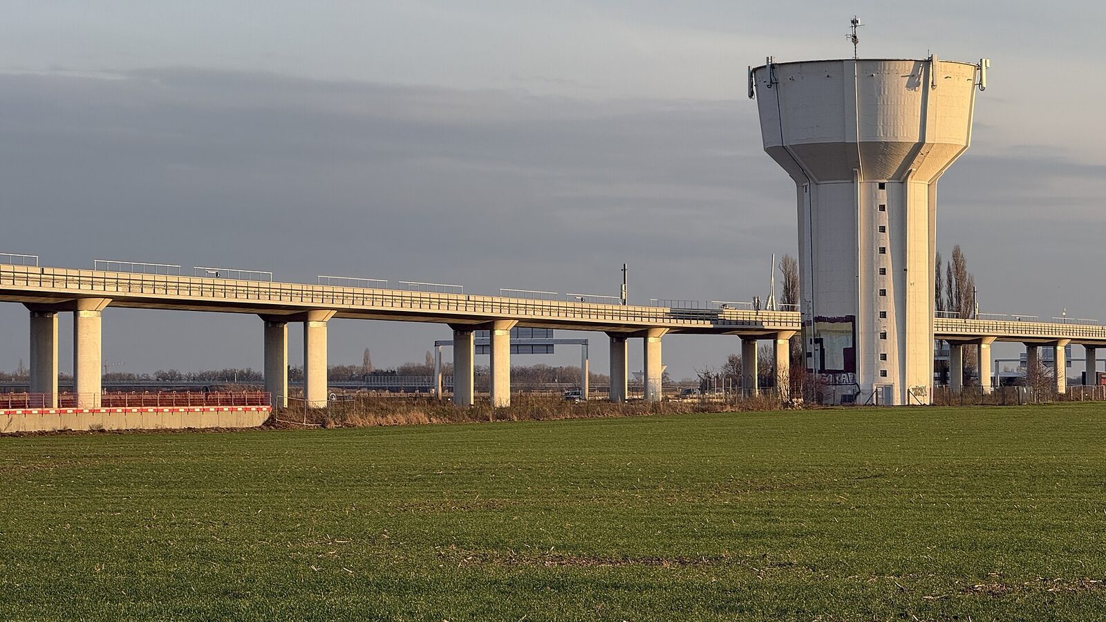 Le long viaduc clair de la ligne 18 traverse les champs du plateau de Saclay, devant un château d’eau